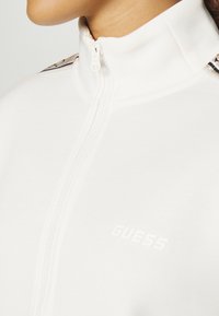 Chaqueta blanca con cremallera y cuello alto, con un sutil logotipo. Textura suave con detalles en contraste en los hombros que aportan un toque distintivo.