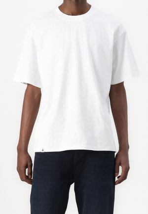 Homme portant un t-shirt blanc uni à manches courtes et un pantalon foncé, debout contre un fond clair avec les bras détendus le long du corps.