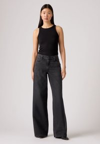 Juodas megztas be rankovių topas derinamas su laisvais, aukšto liemens juodais džinsais. Džinsai turi sagų užsegimą ir šonines kišenes; abu audiniai yra glotniai tekstūruoti.