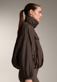 Brown padded jacket with a high collar and elastic cuffs, featuring a front pocket and a loose fit, styled with a smooth texture.