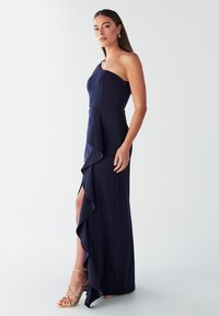 Tumesinine ühe õlaga kleit, millel on vormitud ülakeha, küljerüüsidega ja reie kõrge lõhikuga, valmistatud siledast kangast, kantud kuldsete rihmikõrgete kontsadega.