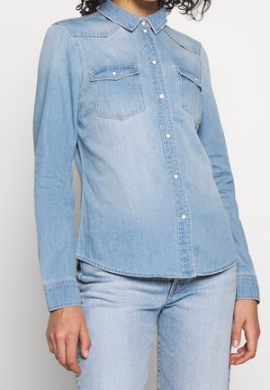 Femme portant une chemise en jean bleu clair à manches longues, boutonnée et avec deux poches poitrine, par-dessus un jean bleu clair.
