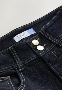 Jeans cu venit larg, de tip high-rise, din denim închis. Prezintă două butoane metalice, detalii cu eticheta albastră și cusături vizibile. Textura este netedă, cu un luciu ușor.