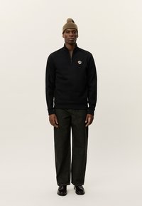Marinblå zip-pullover med ribbade muddar och nederkant, prydd med en liten blomsteremblem; matchas med mörka vida byxor och en beige mössa.