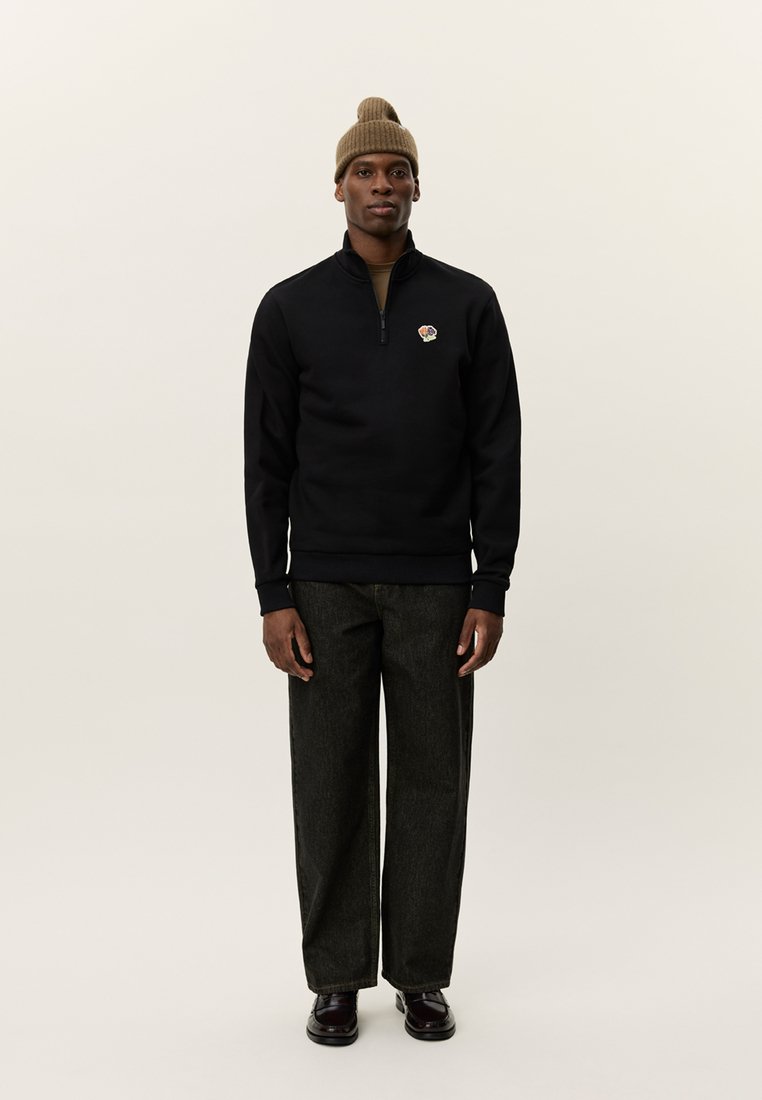 Marinblå zip-pullover med ribbade muddar och nederkant, prydd med en liten blomsteremblem; matchas med mörka vida byxor och en beige mössa.