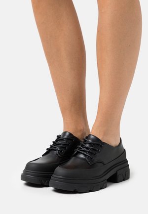 Legs wearing black chunky leather lace-up shoes with thick textured soles, standing on a light background.