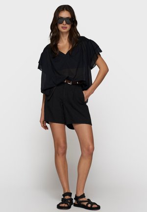 Mujer con gafas de sol negras de gran tamaño, blusa negra holgada, pantalones cortos negros con cinturón y sandalias de cuerda negras, de pie con una mano en el bolsillo.