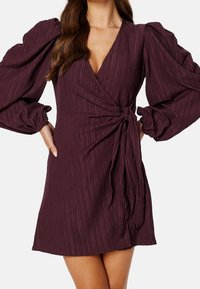Vestido burdeos oscuro con diseño cruzado, que presenta una tela texturizada, voluminosas mangas abullonadas y un acento de cintura atada.