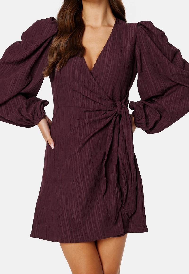 Vestido burdeos oscuro con diseño cruzado, que presenta una tela texturizada, voluminosas mangas abullonadas y un acento de cintura atada.