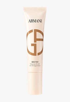 Trousse beige d'Armani Skin Tint avec une finition brillante, présentant un grand logo brun et du texte dans une police simple sur un fond blanc.