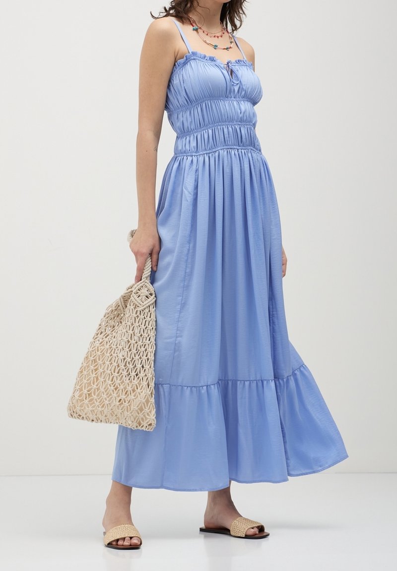 Robe longue bleu clair avec bretelles spaghetti réglables, corsage froncé, ourlet à volants, associée à un sac tressé beige et des sandales slides marron clair.