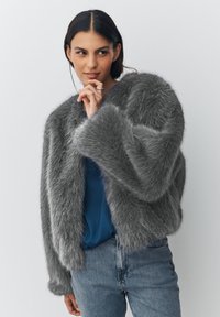 Young woman with dark hair wears a gray faux fur jacket, blue top, and jeans, posing with one hand near her chin against a plain background.