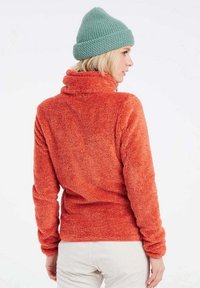 Pullover arancione in pile con colletto alto e maniche lunghe. Texture morbida, aderente in vita, in contrasto con un berretto di maglia blu chiaro.