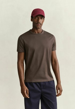 Mann trägt braunes kurzärmeliges T-Shirt, marineblaue Hose und rote Baseballmütze, steht vor einfachem hellem Hintergrund.