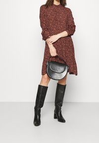 Červené šaty s květinovým vzorem a dlouhými rukávy, doplněné texturovanou černou kabelkou a černými koženými holínkami až po kolena.