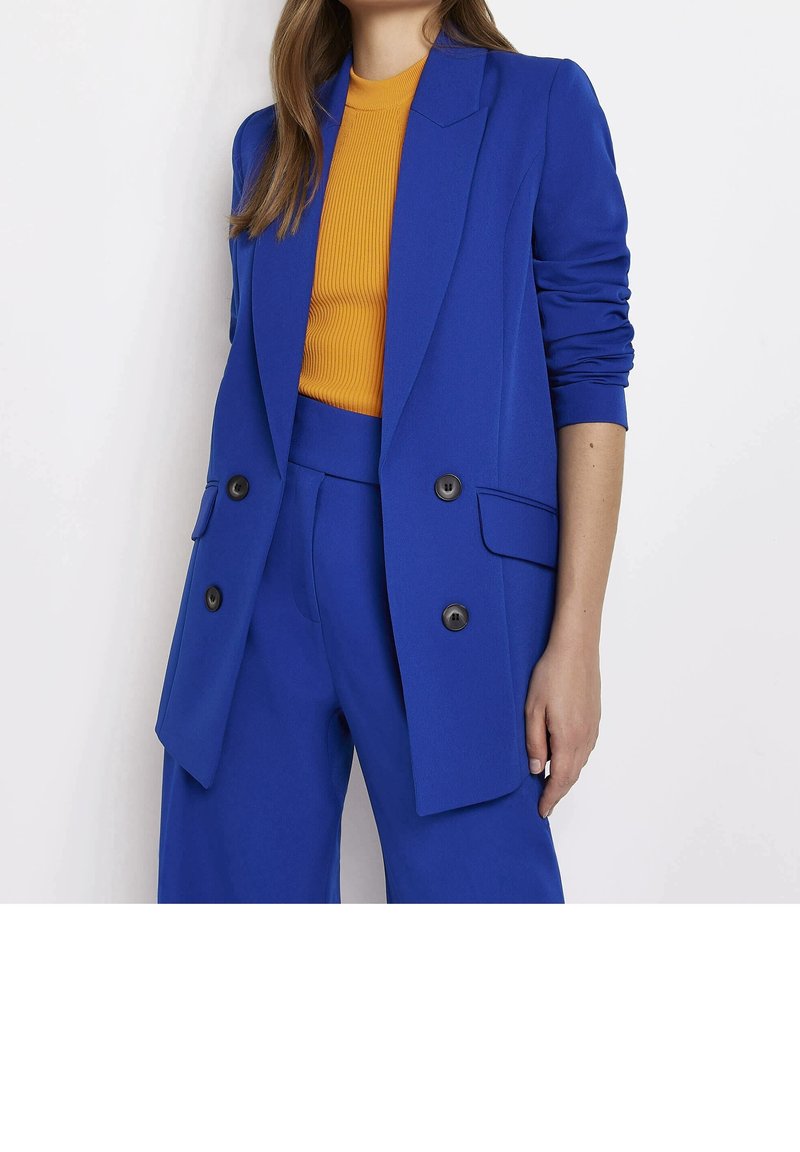 Femme portant un blazer bleu vif à double boutonnage et un pantalon assorti, avec un top côtelé jaune moutarde, debout devant un fond blanc.