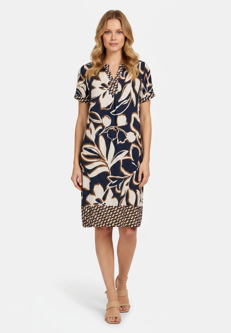 Femme portant une robe mi-longue à manches courtes avec de grands motifs floraux abstraits beige et marron sur un fond sombre.
