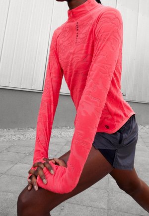 Person strækker benet ud udendørs iført en lys koralfarvet langærmet sportstop og mørkegrå sportsshorts på asfalteret underlag.