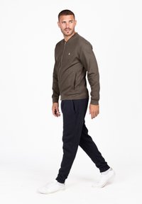 Veste zippée vert olive avec un col rond et des poches latérales, assortie à un pantalon de jogging bleu marine et des baskets blanches.
