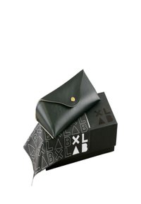 Custodia in pelle nera con una chiusura in ottone, posata su una scatola nera con un motivo geometrico bianco contenente del testo. Include un panno per la pulizia.