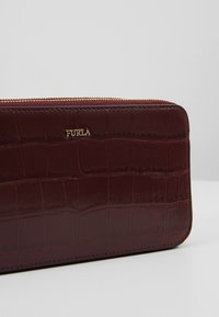 Bordeaux-nahkainen lompakko, jossa on teksturoitu krokotiili-embossattua kuviointia, kultasävytteinen logo ja vetoketjusulku. Suora pinta ja tyylikäs suorakulmainen muoto.