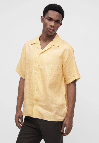 Jeune homme portant une chemise jaune à manches courtes boutonnée et un pantalon sombre, se tenant avec une expression neutre devant un fond gris uni.
