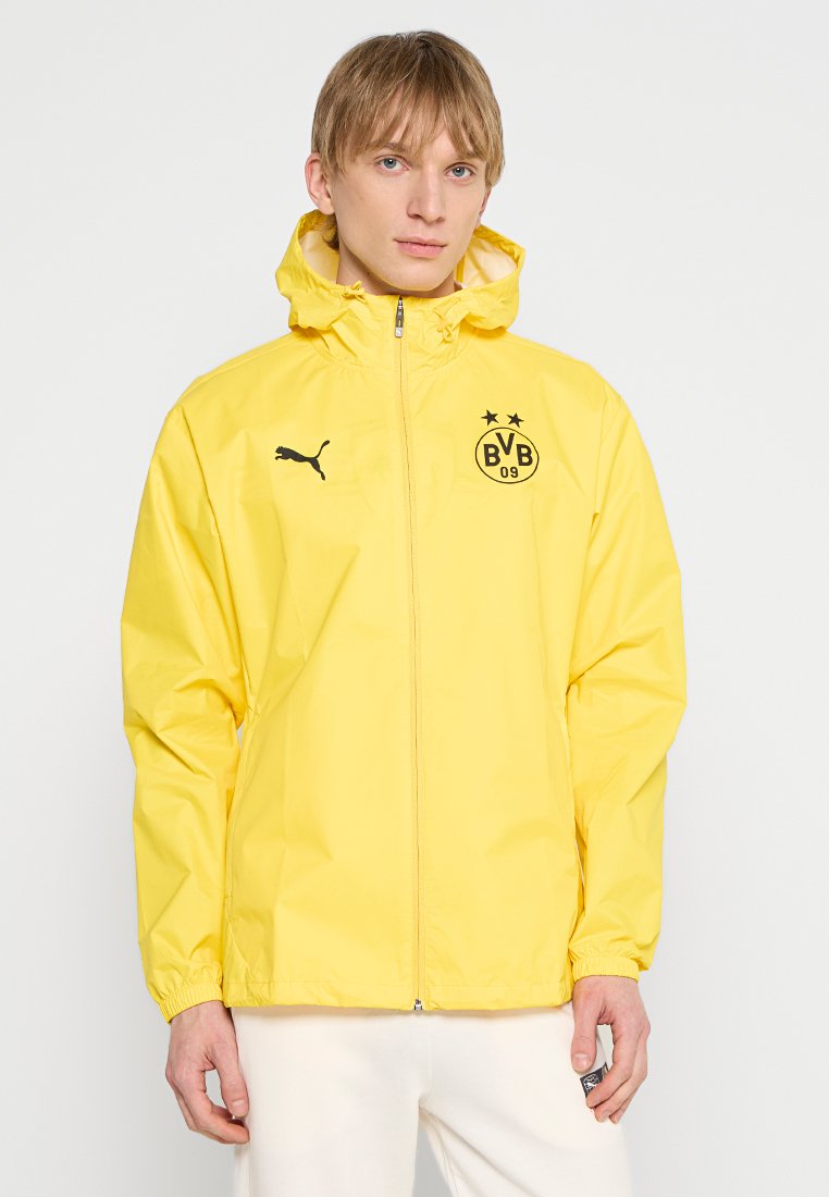 Giacca gialla leggera con zip frontale, polsini elastici e cappuccio. Presenta un logo Puma nero e un emblema della squadra di Dortmund sul petto.