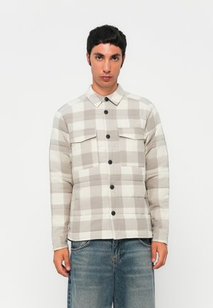 Man wearing beige and white checkered button-up jacket with two chest pockets and blue jeans, standing against a plain white background.