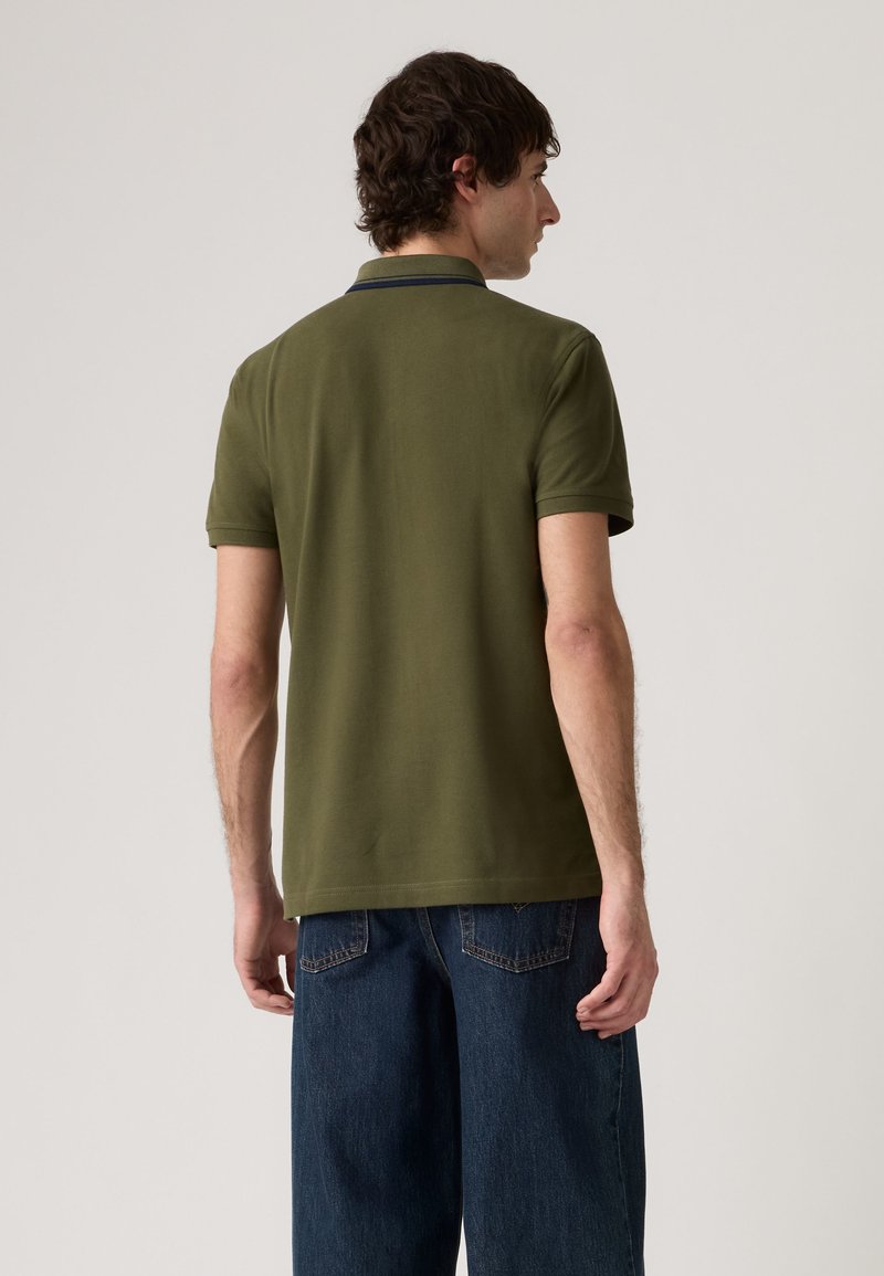 Homme portant un polo manches courtes vert olive et un jean bleu foncé, debout de dos devant un fond clair uni.