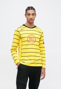Žltá košeľa s dlhým rukávom má čierne horizontálne pruhy, okrúhly oranžový znak v strede a čierny golier. Na pleci je značka Nike.
