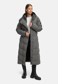 Lång pufferjacka i grått, med kviltad design, huva och knäppning. Bärs över en vit topp och ihop med svarta ankellåga stövlar.