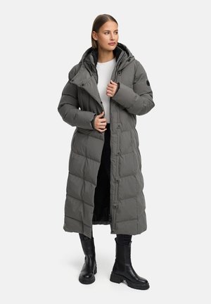 Dlhý puffový kabát v šedé farbe, s prešívaným dizajnom, kapucňou a gombíkovým zapínaním. Nosený cez biele tričko a spárovaný s čiernymi členkovými čižmami.