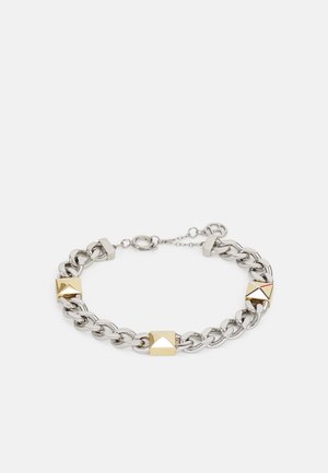 Bracelet en chaîne métallique argentée avec des accents dorés en forme de pyramide, un fermoir à bascule et une longueur adjustable. Texture lisse et brillante.