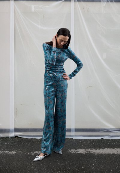 Τοπ σε τσακισμένη υφή σε αποχρώσεις τιρκουάζ, μακρυμάνικο και εφαρμοστό, συνδυασμένο με παντελόνια φαρδιά όψη και με μοτίβο λουλουδιών. Ανάλαφρη γυαλιστερή υφή και ασημένιες λεπτομέρειες στα παπούτσια. Φόντο με κρεμασμένο πλαστικό.