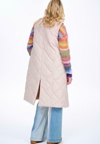 Beżowa pikowana długa kamizelka z bocznymi rozporkami, noszona na wielokolorowy sweter w paski. Luźne dżinsy dopełniają stylizację.