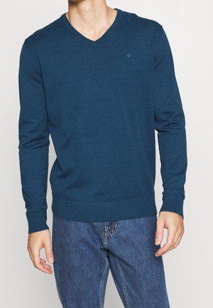 Maglione blu a collo a V in morbido tessuto a maglia, con maniche lunghe e polsini a coste. Dettaglio logo discreto sul petto. Abbinato a jeans di denim.