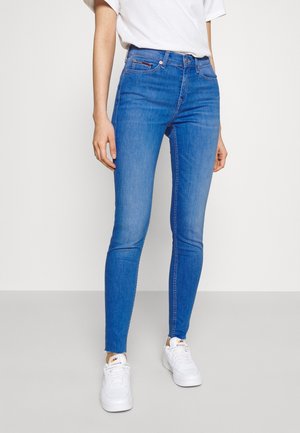Personne portant un jean bleu ajusté, un t-shirt blanc et des baskets blanches, debout sur un sol uni gris clair avec un fond blanc.