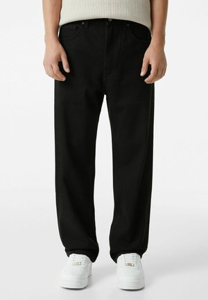 Person trägt lockere schwarze Hose, weiße Sneaker und einen beigen Strickpullover, steht vor einem schlichten weißen Hintergrund.