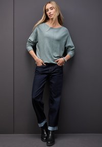Jasnozielony sweter z dzianiny o szerokim dekolcie, w połączeniu z ciemnoniebieskimi dżinsami i czarnymi botkami za kostkę. Modelka stoi na szarym tle.