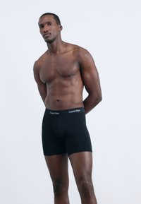 Fekete pamut boxeralsó puha derékpánttal, amelyen a Calvin Klein logó látható, férfi modellel, világos háttér előtt.