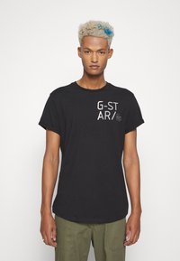 T-shirt de algodão preto com mangas curtas, apresentando texto gráfico branco e logotipo na parte da frente, combinado com calças verde-oliva.