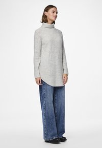 Sieviete stāv, valkājot gaiši pelēku augstu apkaklīti, plašu zilu džinsu bikses un melnus apavus uz vienkārša balta fona.