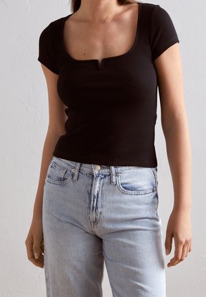 Kvinde iført en sort kortærmet top med firkantet halsudskæring og lyseblå højtaljede jeans mod en ensfarvet væg.