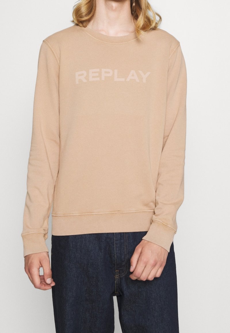 Personne portant un sweat-shirt beige avec le texte "REPLAY" et un jean bleu foncé, debout devant un fond blanc uni.