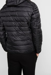 Person iført en sort quilted hættejakke og sorte bukser med hænderne i lommerne, stående foran en ensfarvet lys baggrund.
