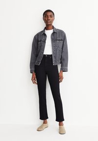 Veste en denim noir à coupe décontractée, portée sur un t-shirt blanc, associée à un jean noir taille haute et des chaussures slip-on beiges.