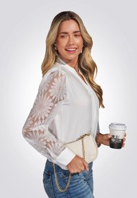 Witte blouse met bloemennetsleeves, lichte denim en een crèmekleurige clutch met een gouden ketting. Houdt een koffiekopje vast met een bedrukt logo.