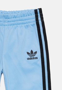 Világoskék sportnadrág, puha anyagból készült, fekete csíkokkal az oldalán és egy fekete Adidas logóval a derékrésznél.