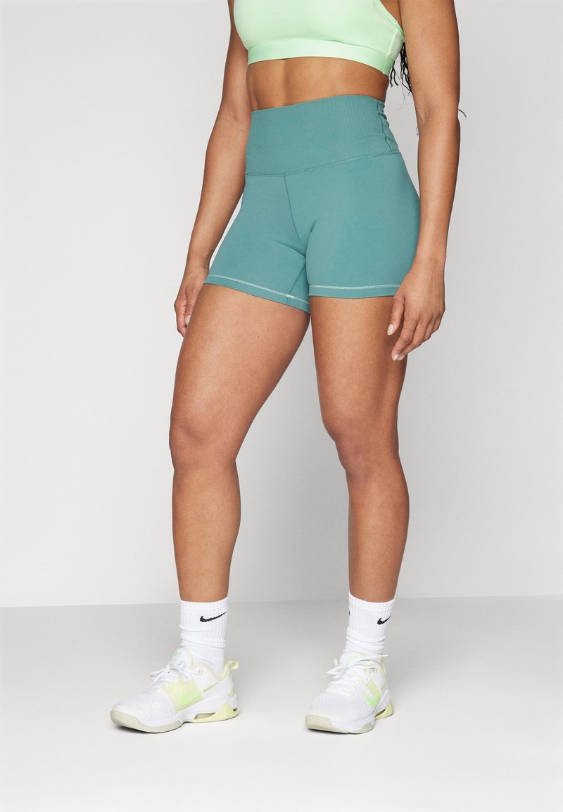 Femme portant un short de sport taille haute couleur sarcelle, un soutien-gorge de sport vert clair, des chaussettes blanches Nike et des baskets blanches, debout devant un fond uni.