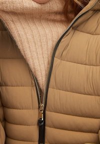 Sandfarvet polstret jakke med quiltet design, hel lynlåslukning og sort kant, lagt over en lys beige ribstrikket sweater.
